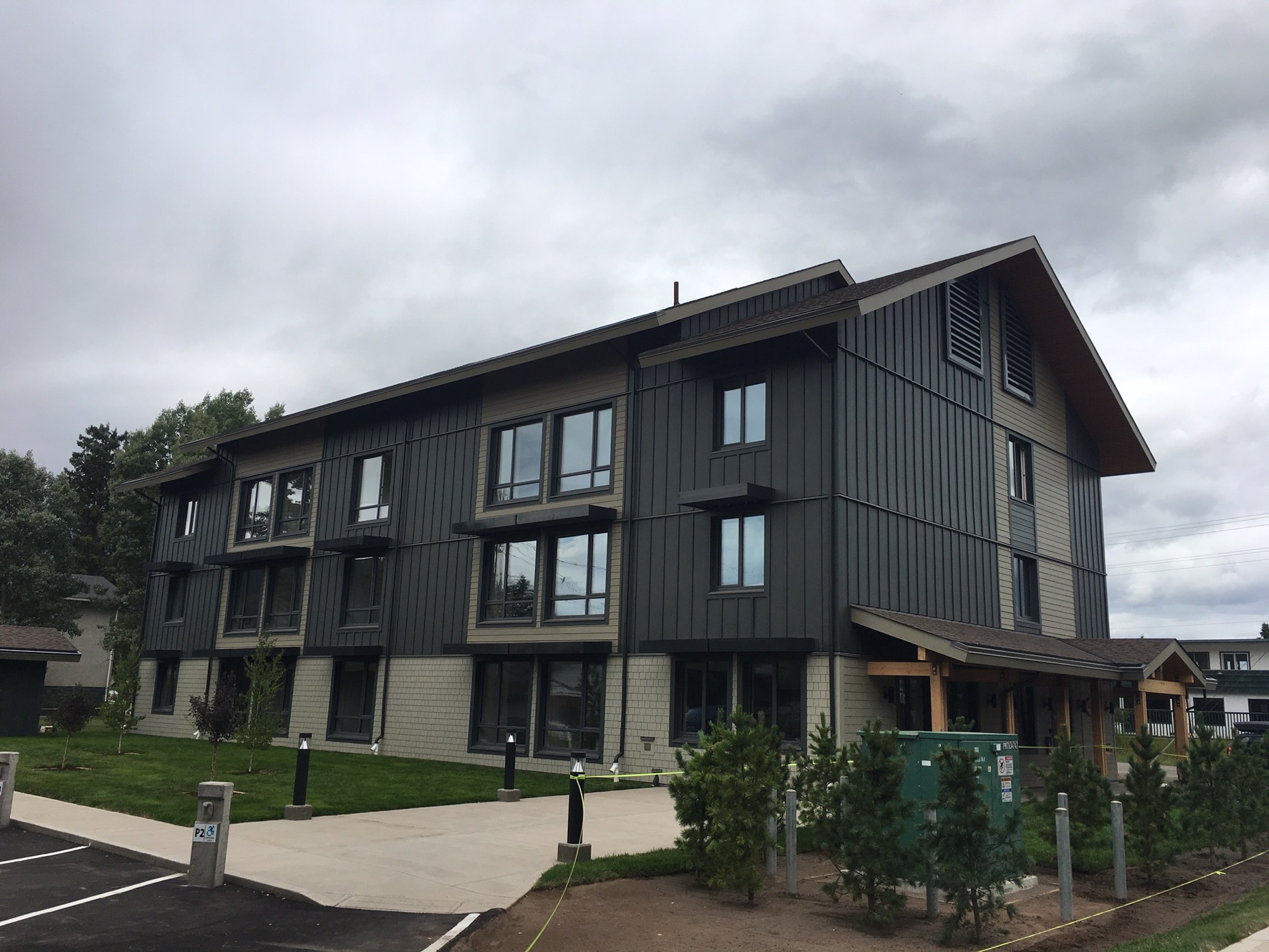 A three-storey timber-clad public building in dark grey and olive, with a covered entrance, a paved accessible parking bay and young planting along the walkway.